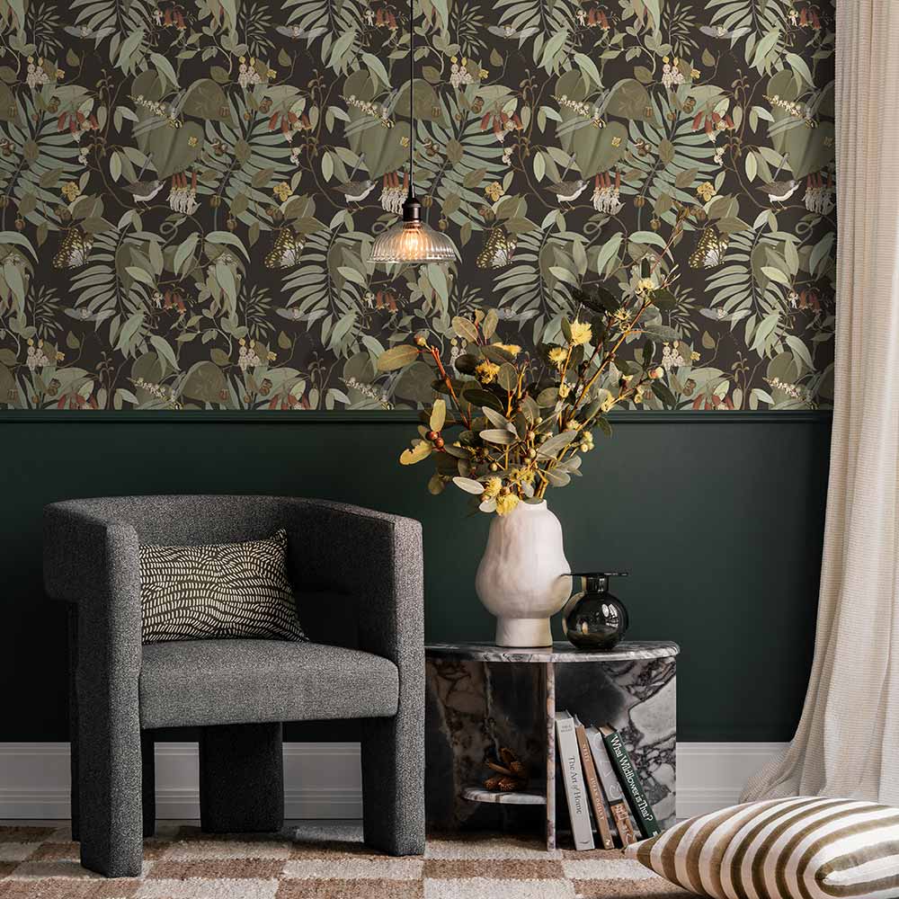 A cozy corner with a dark gray armchair, patterned pillow, marble side table with yellow flowers, and a pendant light. The wall features Urban Road’s Tales of the Bushland Buddies Wallpaper with green tropical leaves above a deep green panel.