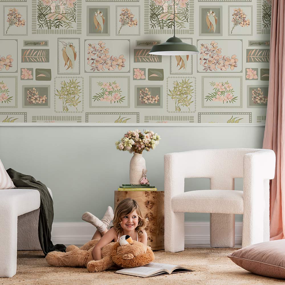 A young child smiles on a beige rug, hugging a teddy bear in a cozy living room featuring Urban Road's Little Bushland Memories Wallpaper, modern white chairs, a wooden side table, and a vase of flowers.