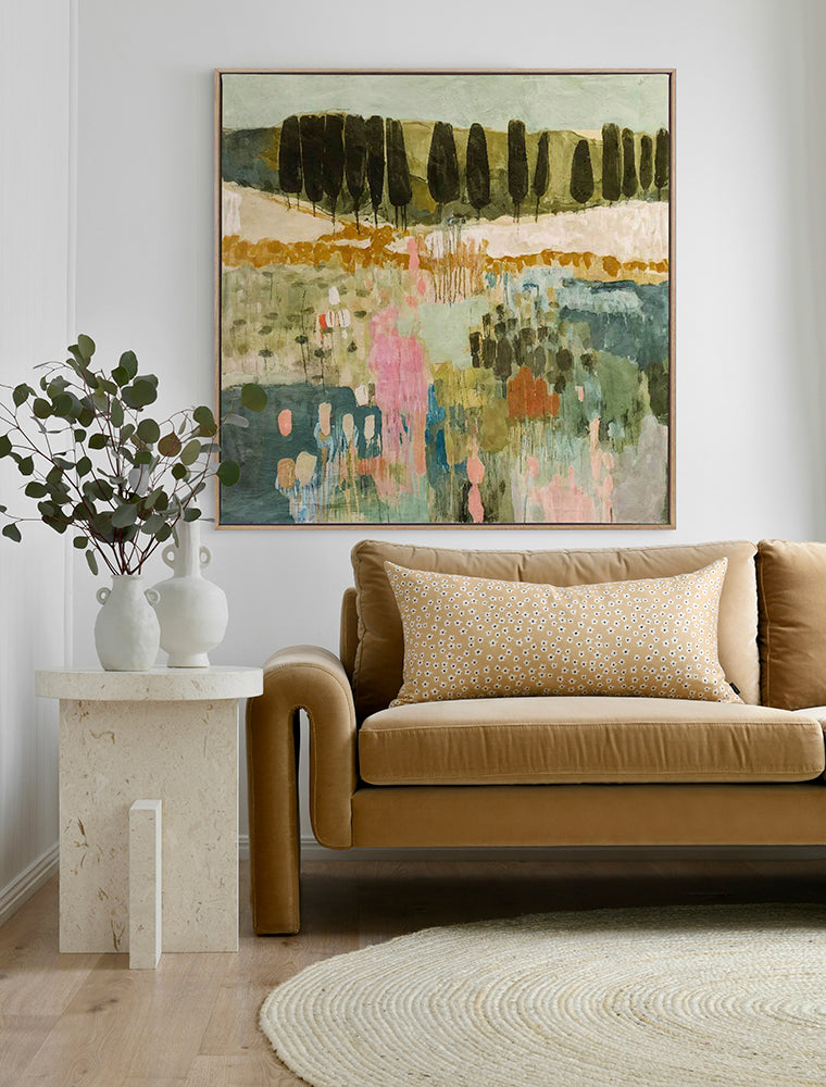 A modern living room with a tan leather sofa, a patterned yellow cushion, a round beige rug, a side table with two white vases and green foliage, and Urban Road's Finding Your Way III - Oak Box Frame Canvas (90x90) on the wall.