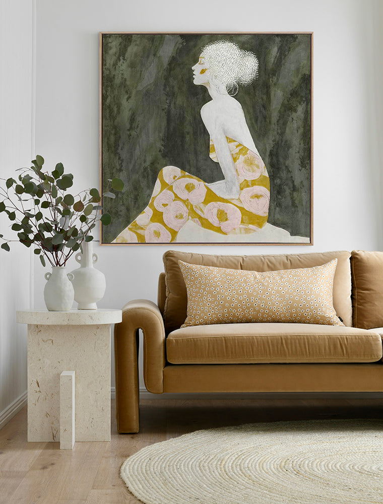 A modern living room with a tan sofa, patterned cushion, round rug, and side table with two white vases and greenery. The wall features Urban Road’s “Solitude in Bloom” oak box frame canvas (90x90) depicting a woman in a floral dress.