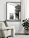 A Rodeo Drive - Black Box Frame Canvas (90x120, Portrait) by Urban Road hangs on a white wall above a plush white chair with a green pillow and a potted plant, creating a modern, inviting atmosphere.