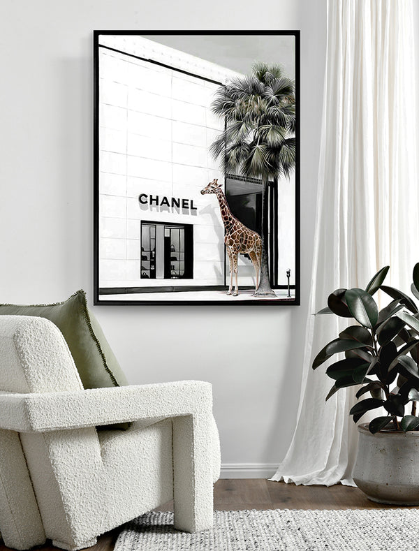 A Rodeo Drive - Black Box Frame Canvas (90x120, Portrait) by Urban Road hangs on a white wall above a plush white chair with a green pillow and a potted plant, creating a modern, inviting atmosphere.