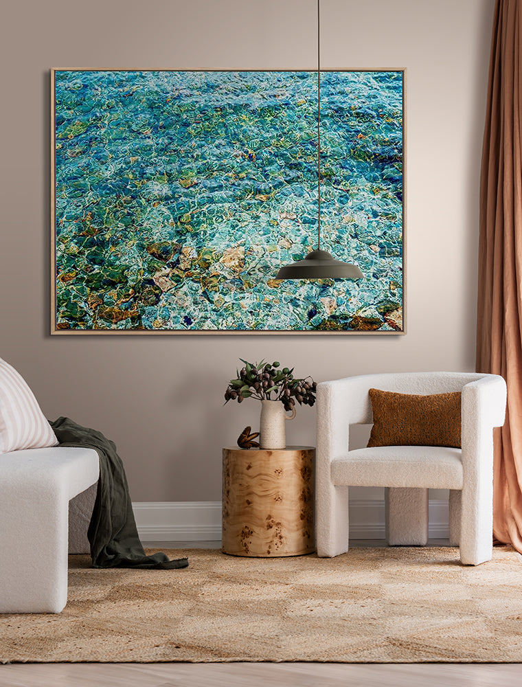 A modern living room features a white armchair, round wooden side table with a vase, tan curtains, a rug, and Urban Road's "The Shallows" oak box frame canvas (90x120 landscape), with tranquil blue-green water artwork above.