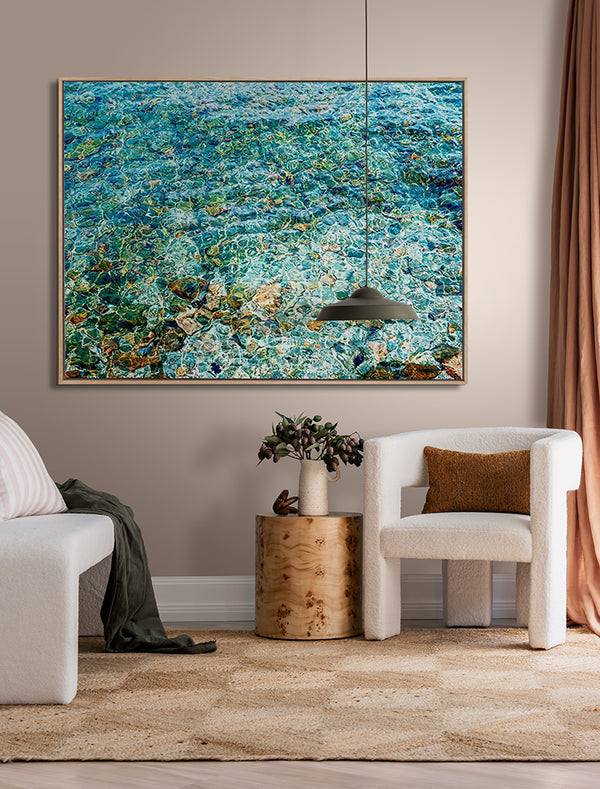 A modern living room features a white armchair, round wooden side table with a vase, tan curtains, a rug, and Urban Road's "The Shallows" oak box frame canvas (90x120 landscape), with tranquil blue-green water artwork above.