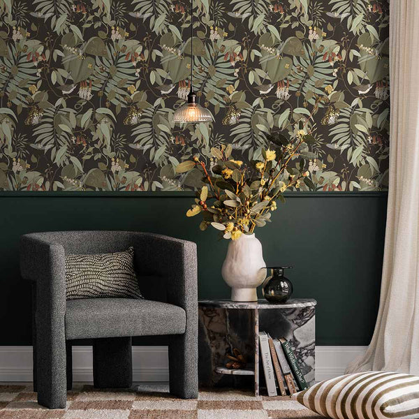 A cozy corner with a dark gray armchair, patterned pillow, marble side table with yellow flowers, and a pendant light. The wall features Urban Road’s Tales of the Bushland Buddies Wallpaper with green tropical leaves above a deep green panel.
