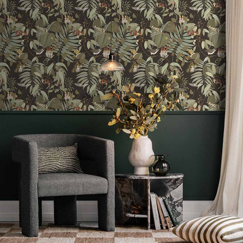 A cozy corner with a dark gray armchair, patterned pillow, marble side table with yellow flowers, and a pendant light. The wall features Urban Road’s Tales of the Bushland Buddies Wallpaper with green tropical leaves above a deep green panel.
