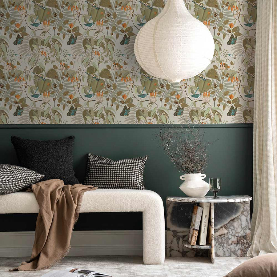 A cozy corner with a dark gray armchair, patterned pillow, marble side table with yellow flowers, and a pendant light. The wall features Urban Road’s Tales of the Bushland Buddies Wallpaper with green tropical leaves above a deep green panel.
