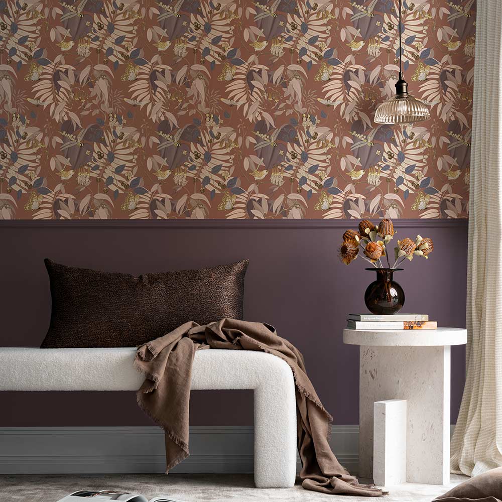 A cozy corner with a dark gray armchair, patterned pillow, marble side table with yellow flowers, and a pendant light. The wall features Urban Road’s Tales of the Bushland Buddies Wallpaper with green tropical leaves above a deep green panel.
