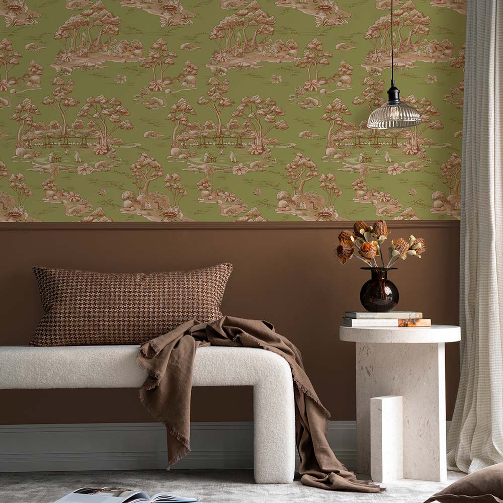 A cozy living room with a cream sofa, striped pillow, and brown blanket sits beside a table with vases and two bear figurines. A round pendant light hangs above, while Urban Road's Bush Baby Village Toile Wallpaper charms the brown wall.
