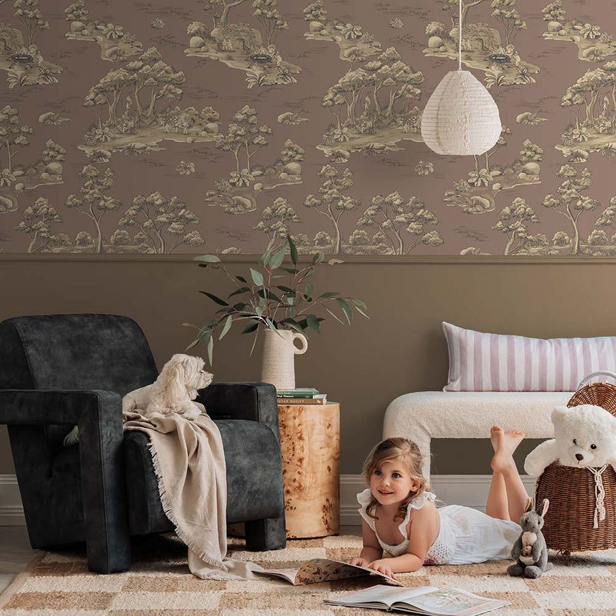 A cozy living room with a cream sofa, striped pillow, and brown blanket sits beside a table with vases and two bear figurines. A round pendant light hangs above, while Urban Road's Bush Baby Village Toile Wallpaper charms the brown wall.
