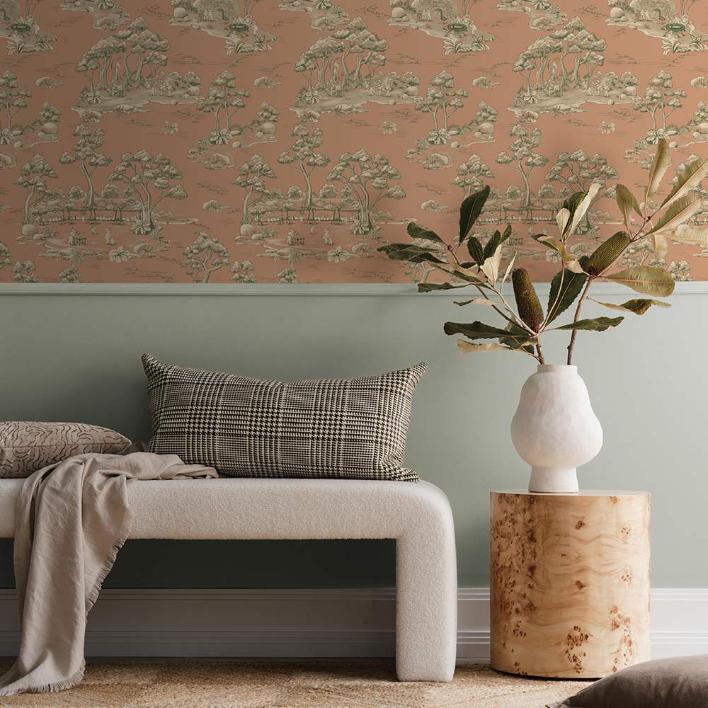 A cozy living room with a cream sofa, striped pillow, and brown blanket sits beside a table with vases and two bear figurines. A round pendant light hangs above, while Urban Road's Bush Baby Village Toile Wallpaper charms the brown wall.
