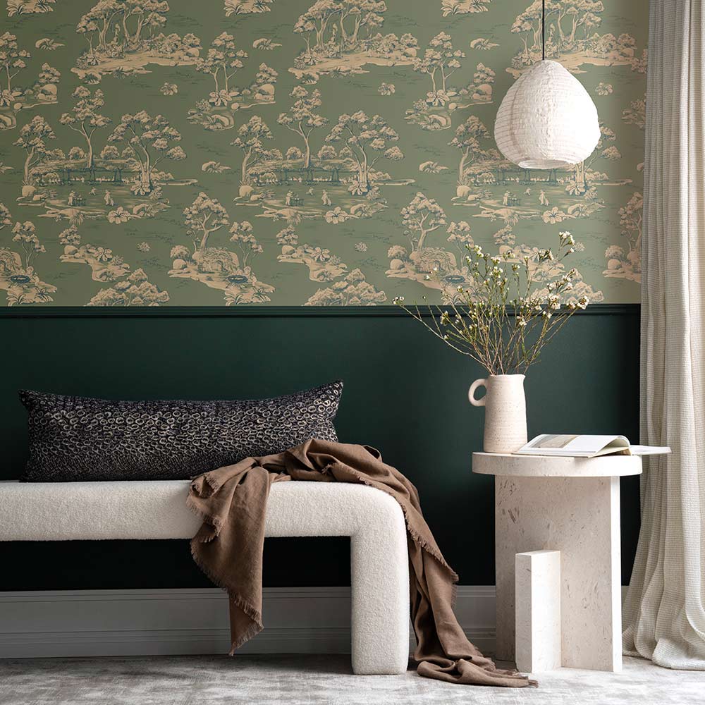 A cozy living room with a cream sofa, striped pillow, and brown blanket sits beside a table with vases and two bear figurines. A round pendant light hangs above, while Urban Road's Bush Baby Village Toile Wallpaper charms the brown wall.
