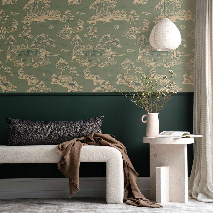 A cozy living room with a cream sofa, striped pillow, and brown blanket sits beside a table with vases and two bear figurines. A round pendant light hangs above, while Urban Road's Bush Baby Village Toile Wallpaper charms the brown wall.
