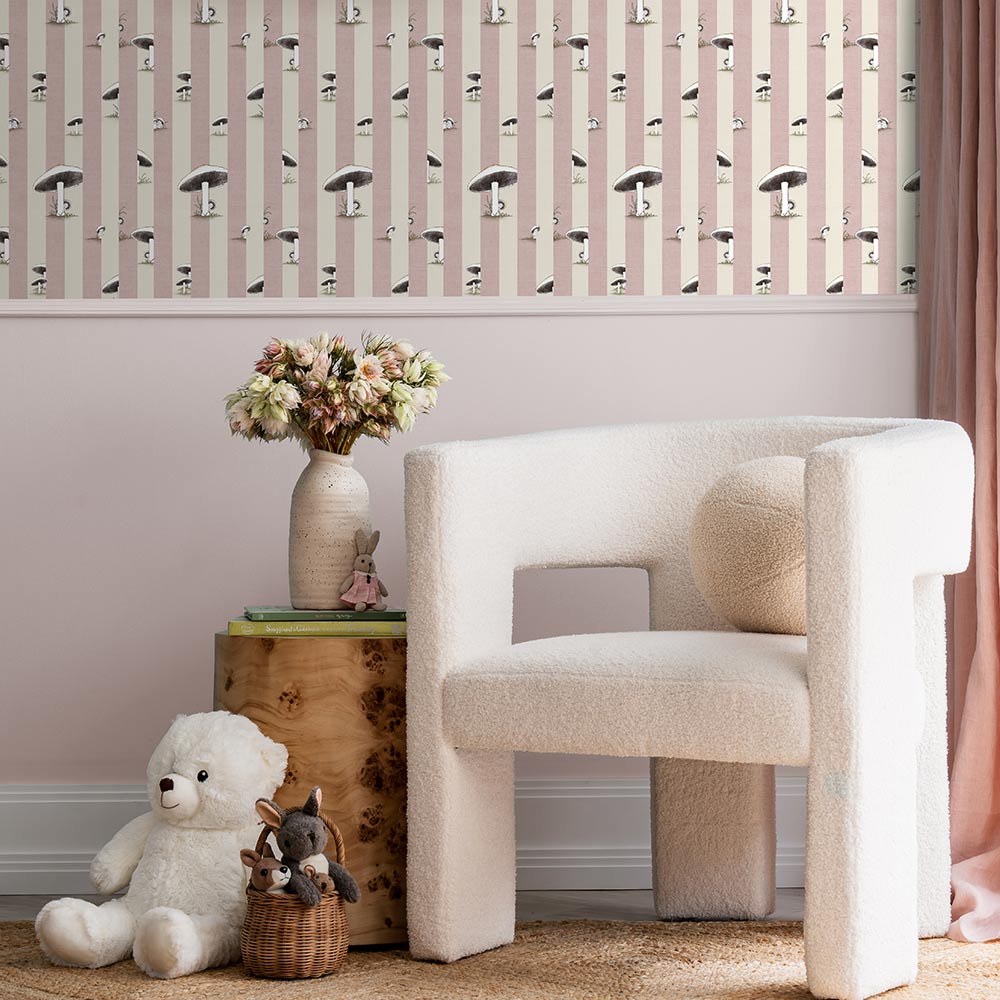 A cozy room features a white plush chair, wooden side table with flowers, two stuffed animals, pink curtains, and Urban Road’s Toadstool Peakaboo Light Wallpaper with vertical stripes and abstract faces. A green pendant light hangs overhead.

