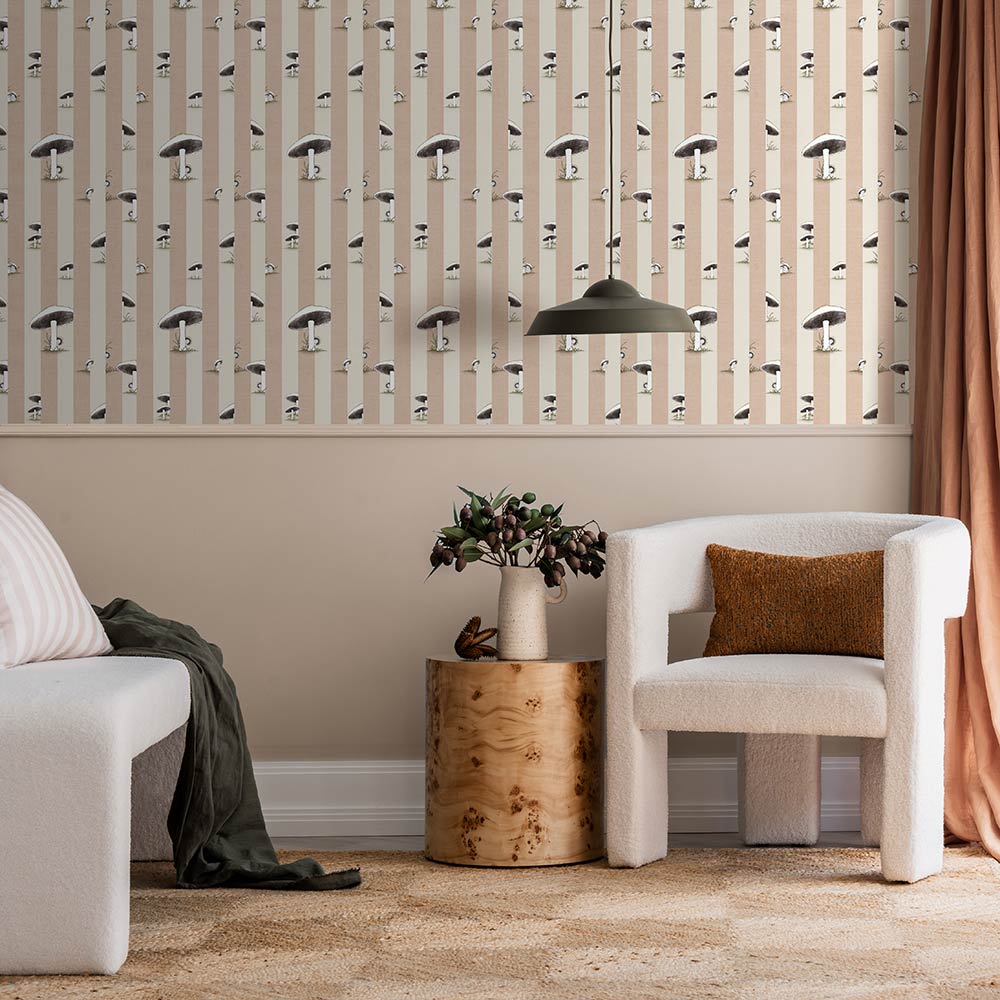 A cozy room features a white plush chair, wooden side table with flowers, two stuffed animals, pink curtains, and Urban Road’s Toadstool Peakaboo Light Wallpaper with vertical stripes and abstract faces. A green pendant light hangs overhead.
