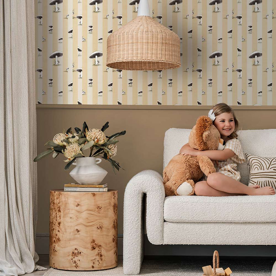 A cozy room features a white plush chair, wooden side table with flowers, two stuffed animals, pink curtains, and Urban Road’s Toadstool Peakaboo Light Wallpaper with vertical stripes and abstract faces. A green pendant light hangs overhead.
