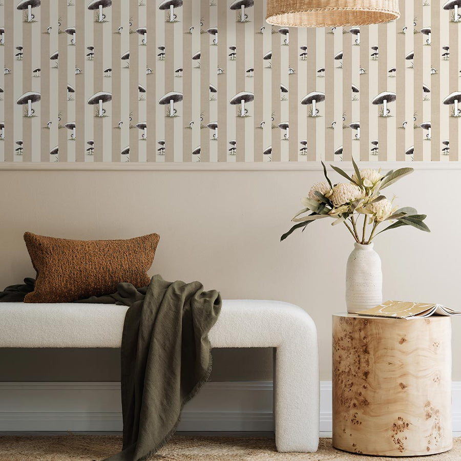 A cozy room features a white plush chair, wooden side table with flowers, two stuffed animals, pink curtains, and Urban Road’s Toadstool Peakaboo Light Wallpaper with vertical stripes and abstract faces. A green pendant light hangs overhead.
