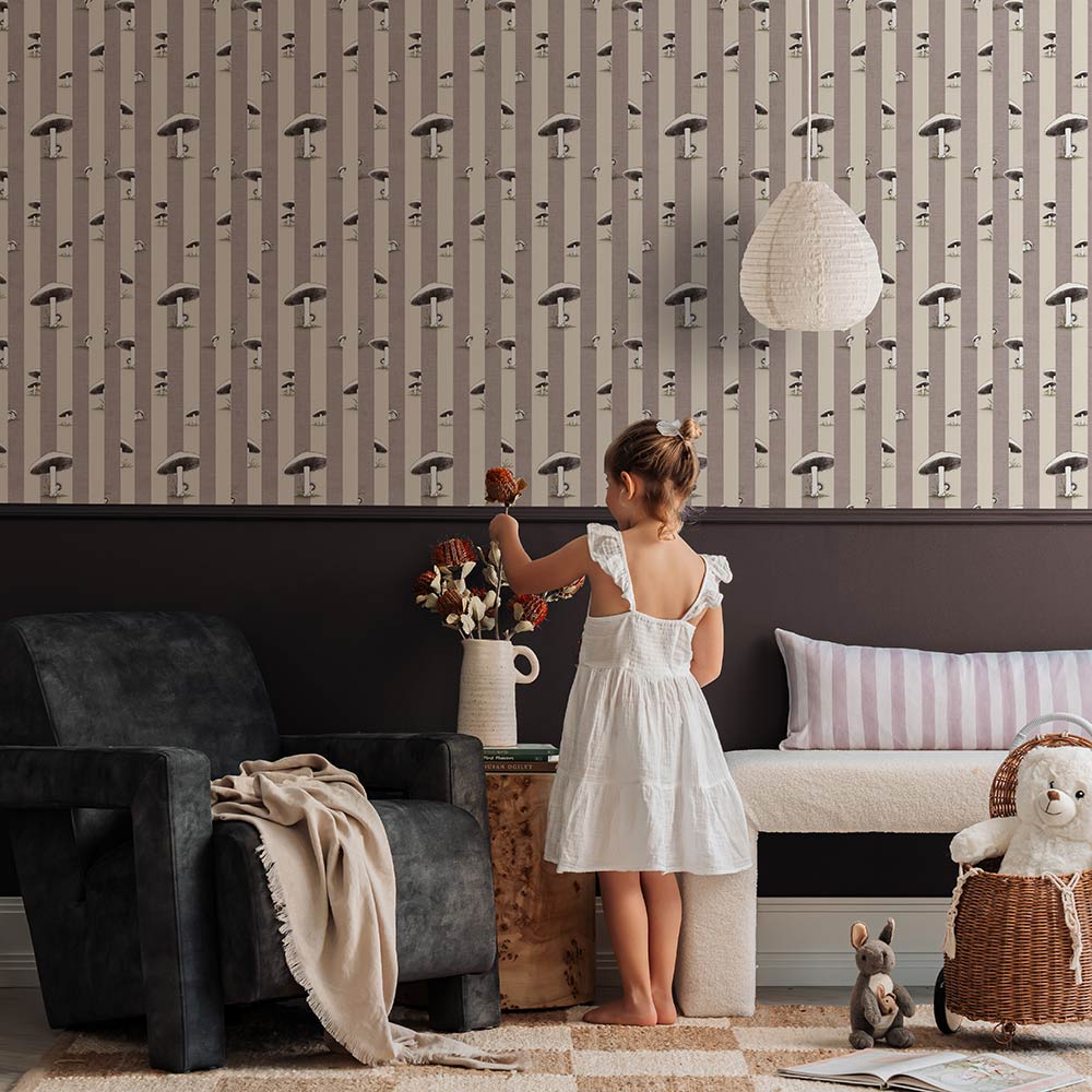 A cozy room features a white plush chair, wooden side table with flowers, two stuffed animals, pink curtains, and Urban Road’s Toadstool Peakaboo Light Wallpaper with vertical stripes and abstract faces. A green pendant light hangs overhead.
