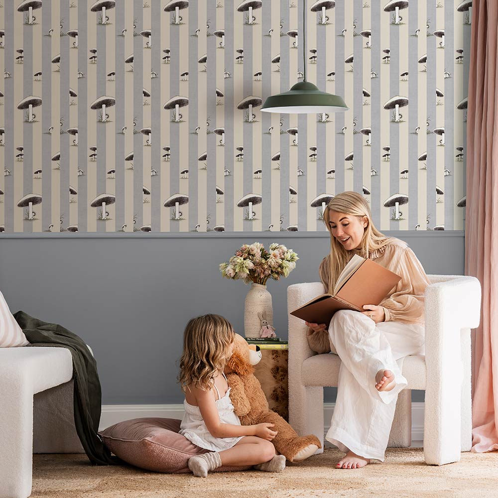 A cozy room features a white plush chair, wooden side table with flowers, two stuffed animals, pink curtains, and Urban Road’s Toadstool Peakaboo Light Wallpaper with vertical stripes and abstract faces. A green pendant light hangs overhead.
