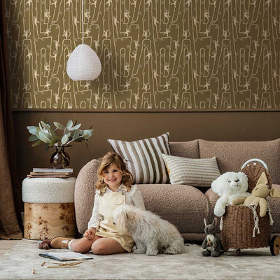 A young girl sits on a rug in a modern living room with a fluffy dog, plush toys, and cushions, all set against the Urban Road Giggly Bush Babies Wallpaper in soft olive green, adding a playful touch to the space.
