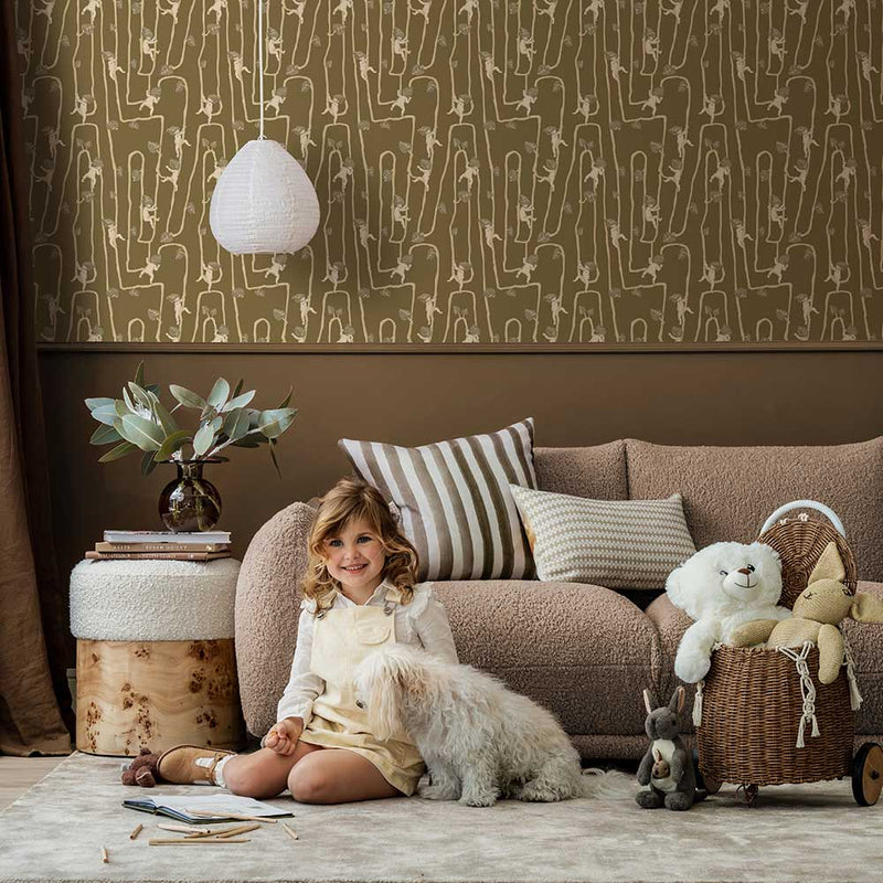 A young girl sits on a rug in a modern living room with a fluffy dog, plush toys, and cushions, all set against the Urban Road Giggly Bush Babies Wallpaper in soft olive green, adding a playful touch to the space.
