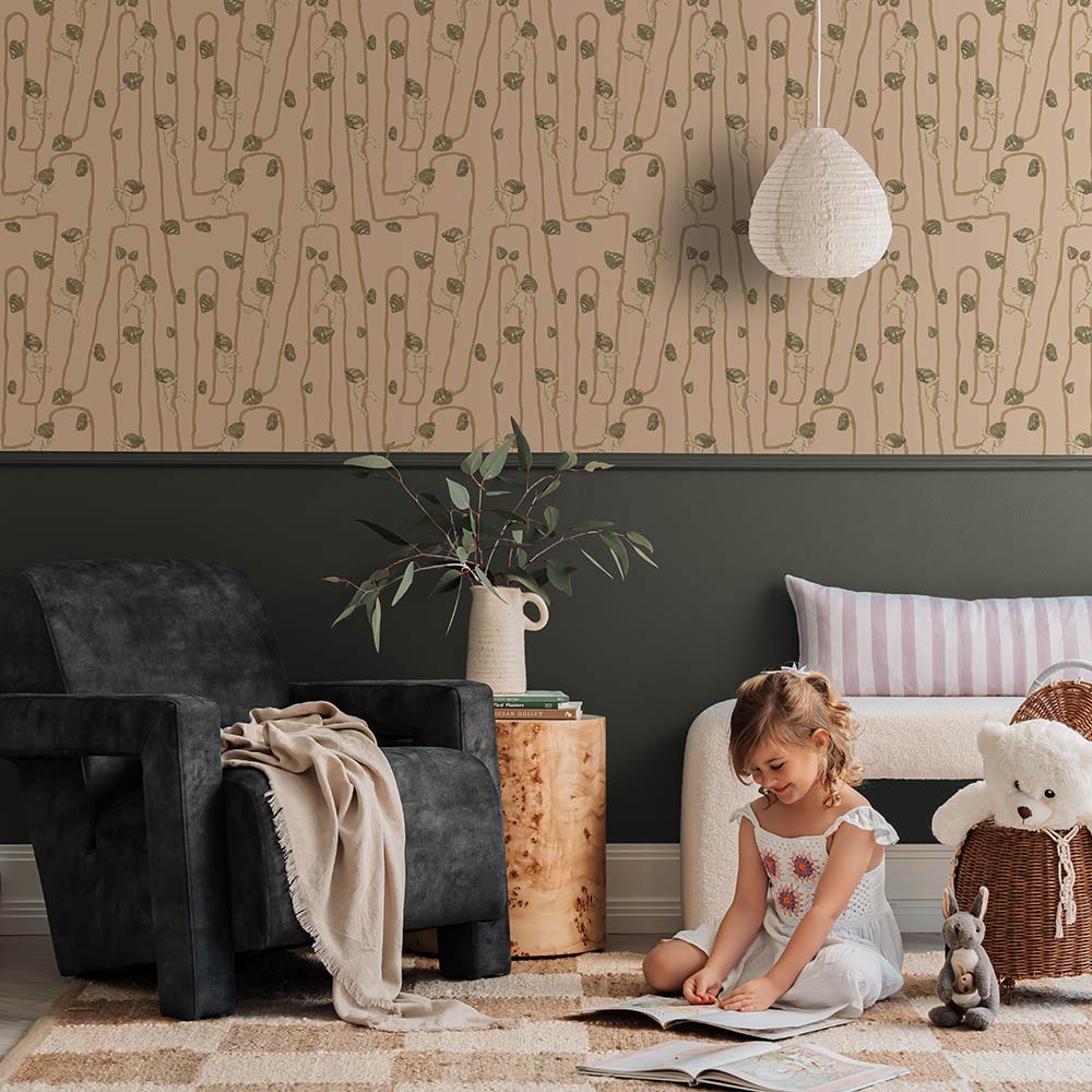 A young girl sits on a rug in a modern living room with a fluffy dog, plush toys, and cushions, all set against the Urban Road Giggly Bush Babies Wallpaper in soft olive green, adding a playful touch to the space.
