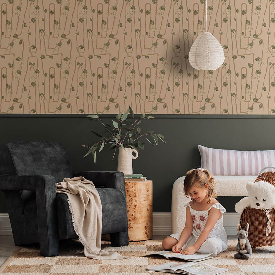 A young girl sits on a rug in a modern living room with a fluffy dog, plush toys, and cushions, all set against the Urban Road Giggly Bush Babies Wallpaper in soft olive green, adding a playful touch to the space.
