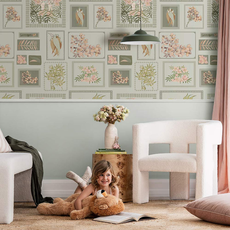 A young child smiles on a beige rug, hugging a teddy bear in a cozy living room featuring Urban Road's Little Bushland Memories Wallpaper, modern white chairs, a wooden side table, and a vase of flowers.
