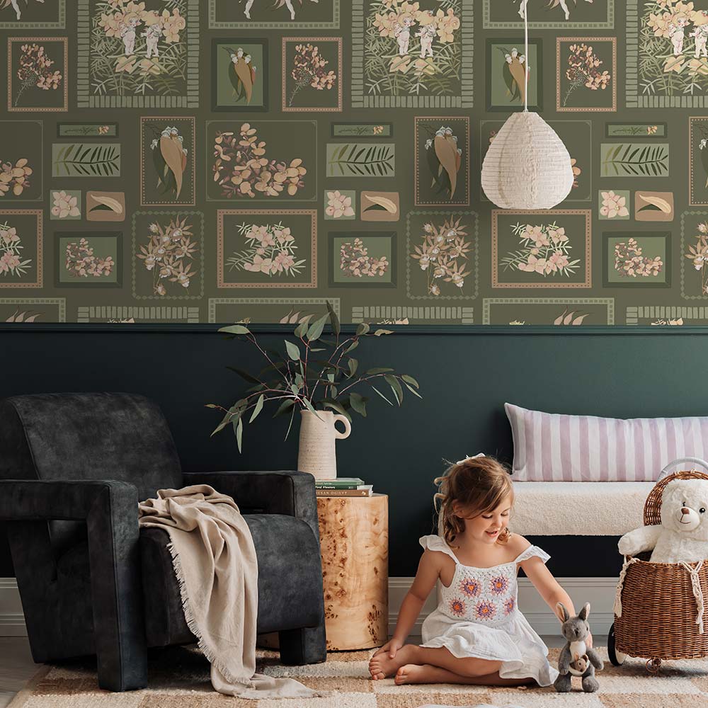 A young child smiles on a beige rug, hugging a teddy bear in a cozy living room featuring Urban Road's Little Bushland Memories Wallpaper, modern white chairs, a wooden side table, and a vase of flowers.
