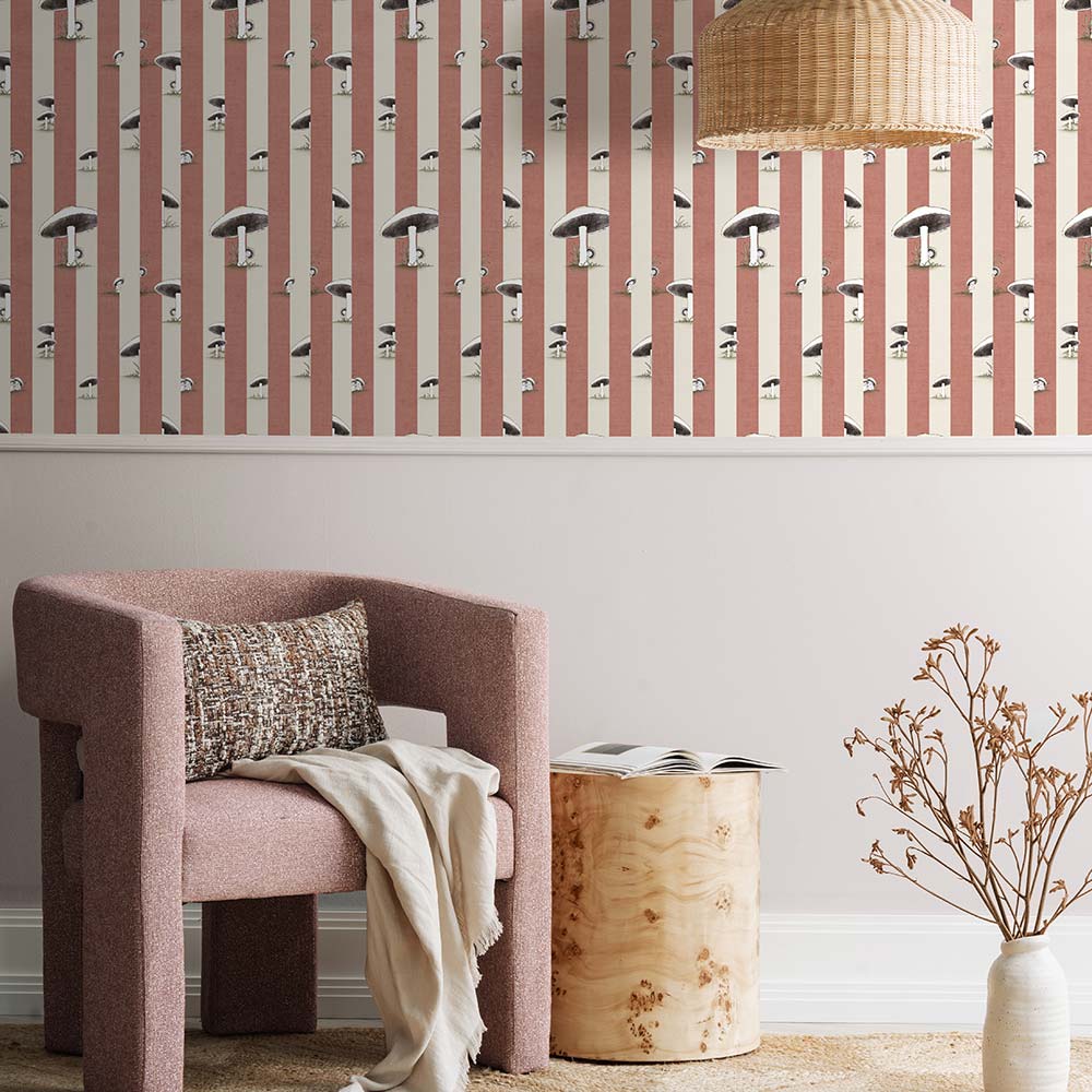 A cozy pink armchair with a cushion sits by a wooden side table and dried flowers, against a wall adorned with Urban Road’s Toadstool Peakaboo Dark Wallpaper, beneath a woven pendant light.
