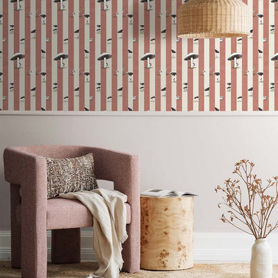A cozy pink armchair with a cushion sits by a wooden side table and dried flowers, against a wall adorned with Urban Road’s Toadstool Peakaboo Dark Wallpaper, beneath a woven pendant light.
