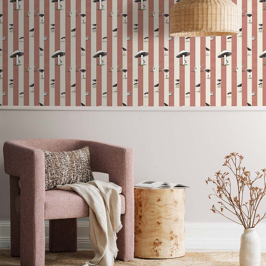 A cozy pink armchair with a cushion sits by a wooden side table and dried flowers, against a wall adorned with Urban Road’s Toadstool Peakaboo Dark Wallpaper, beneath a woven pendant light.
