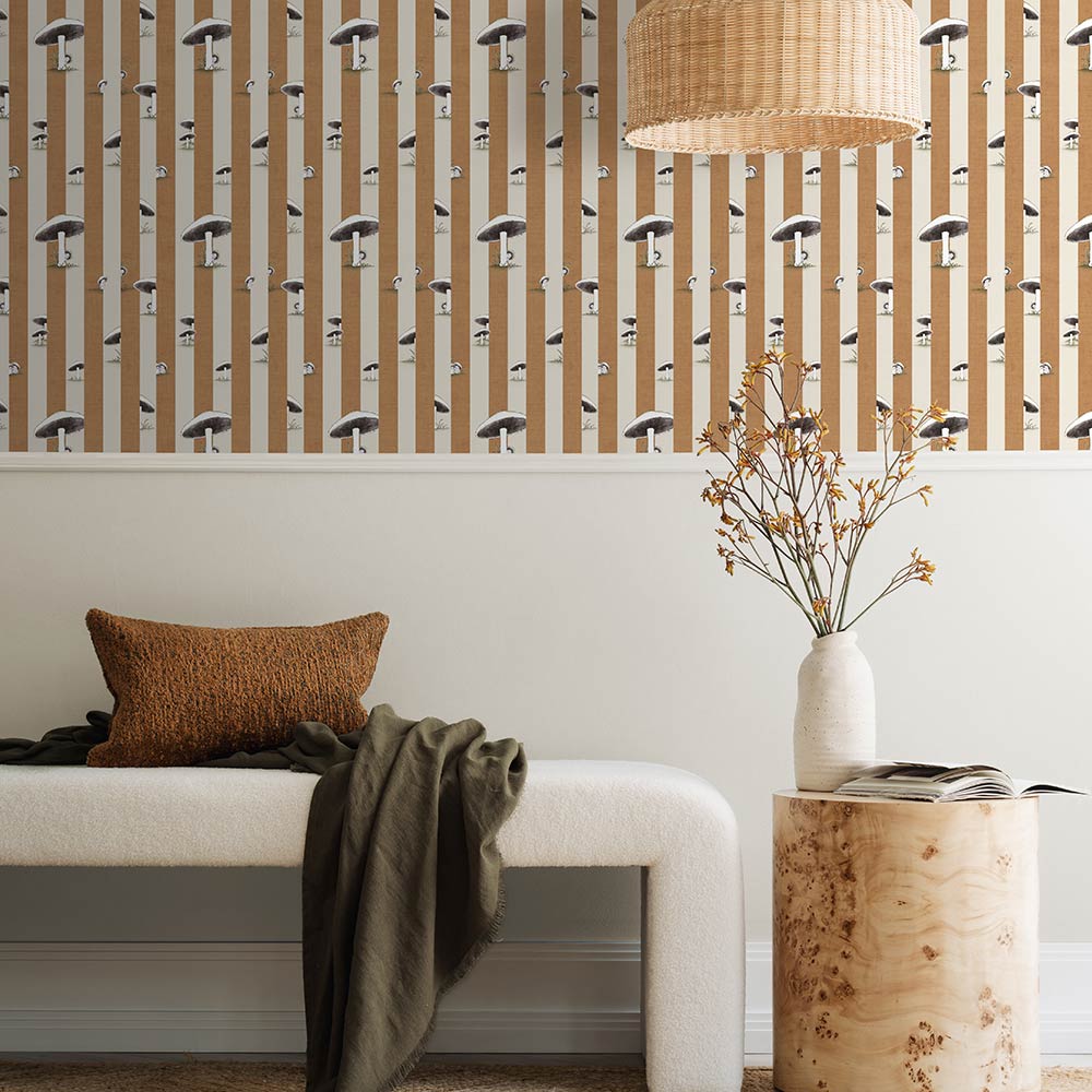 A cozy pink armchair with a cushion sits by a wooden side table and dried flowers, against a wall adorned with Urban Road’s Toadstool Peakaboo Dark Wallpaper, beneath a woven pendant light.

