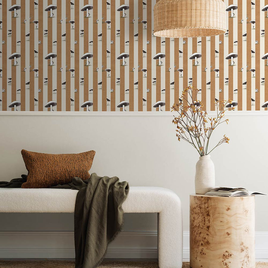 A cozy pink armchair with a cushion sits by a wooden side table and dried flowers, against a wall adorned with Urban Road’s Toadstool Peakaboo Dark Wallpaper, beneath a woven pendant light.
