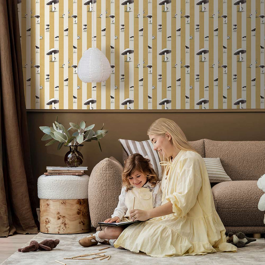 A cozy pink armchair with a cushion sits by a wooden side table and dried flowers, against a wall adorned with Urban Road’s Toadstool Peakaboo Dark Wallpaper, beneath a woven pendant light.

