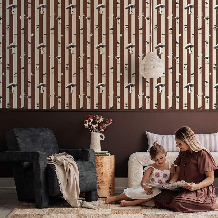 A cozy pink armchair with a cushion sits by a wooden side table and dried flowers, against a wall adorned with Urban Road’s Toadstool Peakaboo Dark Wallpaper, beneath a woven pendant light.
