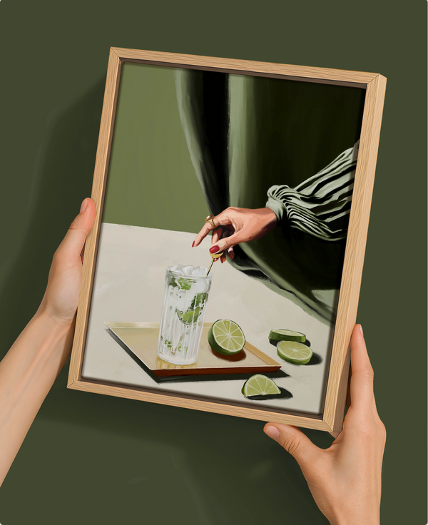 Two hands hold the Urban Road Mojito - Oak Box Frame Canvas (22.5x30, Portrait), featuring a hand stirring a mojito with lime slices before a green curtain. The framed art rests against a dark green background, ideal for modern art lovers.