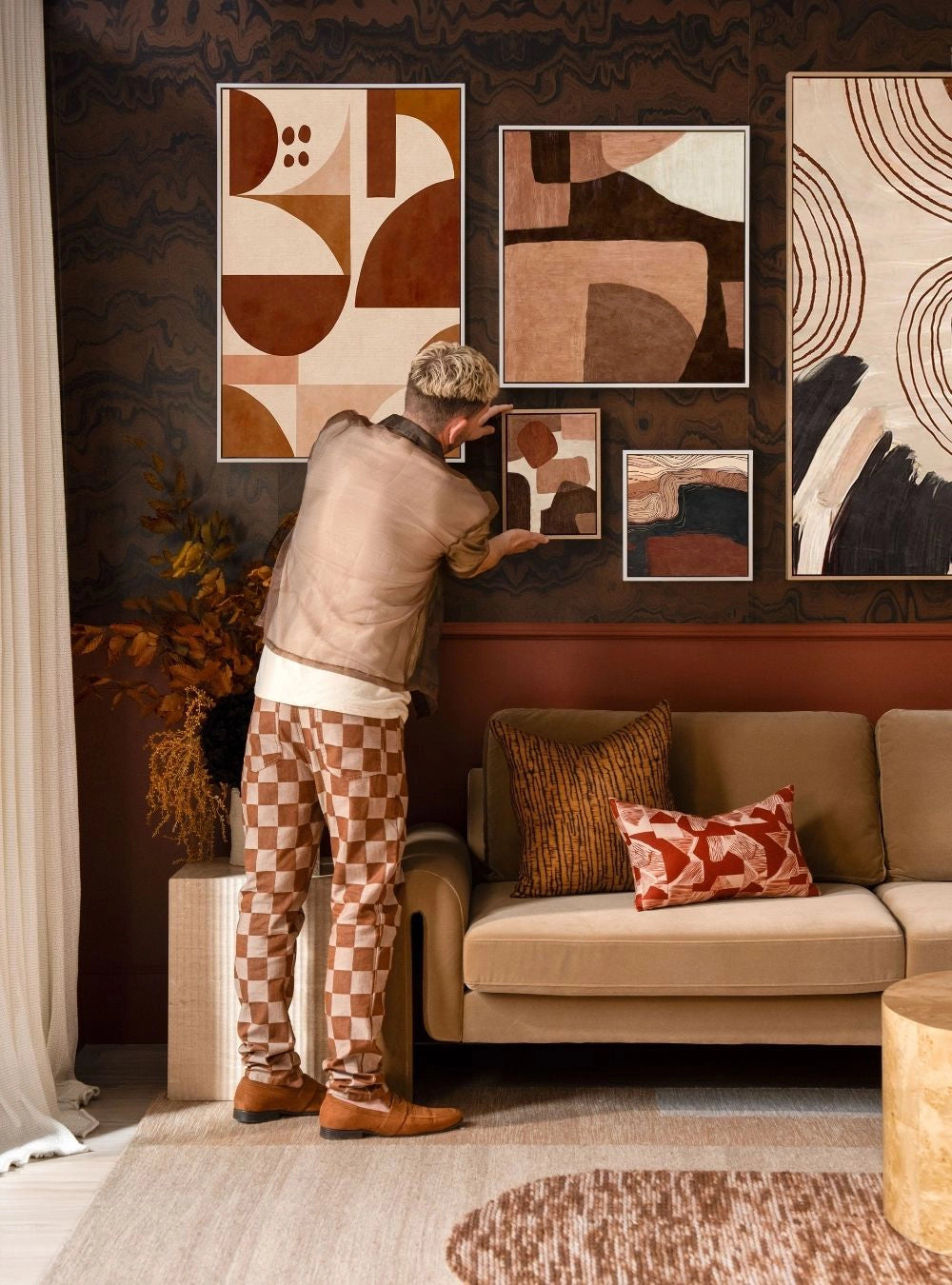 Tim Neve adjusting framed abstract art on a dark brown gallery wall above a beige couch with patterned cushions.