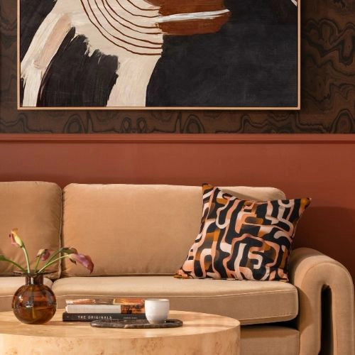 Close-up of a beige couch with a bold, abstract cushion and a round wood table in a warm brown-toned room.