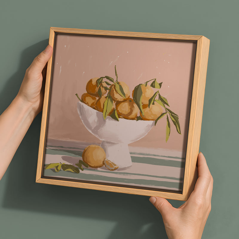 Hands hold the Urban Road Bowl of Oranges - Oak Box Frame Canvas - 30x30, featuring a white bowl with oranges and green leaves on a striped surface against a pink background—an artwork that adds vibrancy to any space.