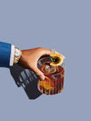A hand with a watch holds a glass of dark cocktail with ice, lemon twist, and cherry against a blue-gray backdrop—Urban Road’s Old Fashioned Canvas Art Print is ideal for modern spaces or the On Vacation Collection.