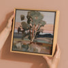 Two hands hold Urban Road's "The Old Gum Tree" 30x30 oak box frame canvas, featuring a single tree in a muted landscape with a pink sky—ideal as Australian outback wall art.