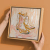 A person holds Urban Roadâ€™s "Cowboy Boots at Sunset" canvas (30x30, oak box frame) against a peach wall. Its expressive beige, brown, orange, and pink brushstrokes add perfect Western style to any space.
