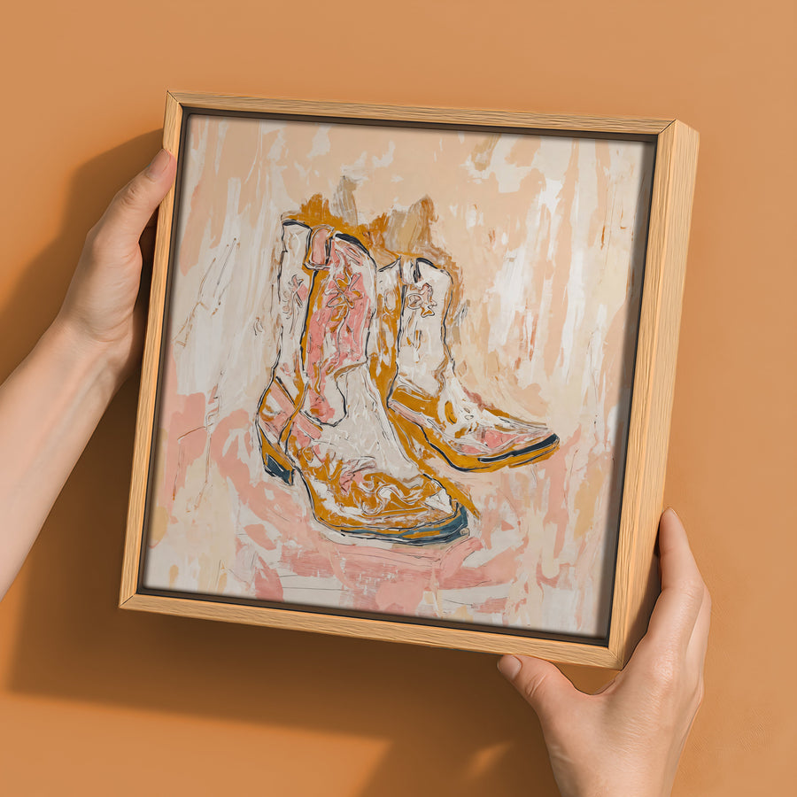 A person holds Urban Roadâ€™s "Cowboy Boots at Sunset" canvas (30x30, oak box frame) against a peach wall. Its expressive beige, brown, orange, and pink brushstrokes add perfect Western style to any space.