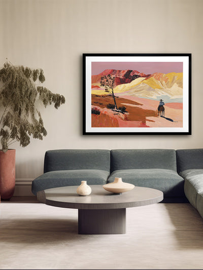 A modern living room features a curved gray sofa, round coffee table, two small vases, a potted plant, and the Rogue Terrain Framed Art Print by Urban Road depicting a Western landscape with mountains and a cowboy on horseback.
