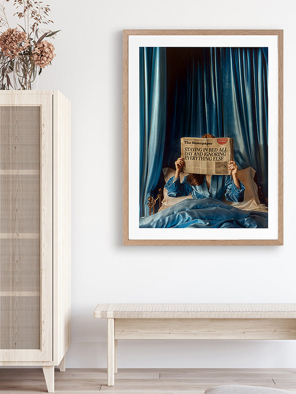 A minimalist room with light wood furniture features the Morning Gossip Framed Art Print by Urban Road, showing a person in bed with a newspaper and blue curtains. Dried flowers sit atop a cabinet on the left.