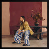 A woman in yellow seated against a moody blue and red backdrop, wearing bold striped pants, from the Lounging Ladies II collection by Urban Road.