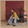 A woman in yellow seated against a moody blue and red backdrop, wearing bold striped pants, from the Lounging Ladies II collection by Urban Road.