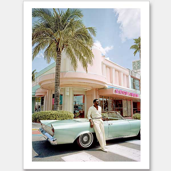 A man in a cream suit leans on a vintage blue convertible before a pastel pink Art Deco building—captured in the "Havana Days Unframed Art Print" by Shaynna Blaze x Urban Road, ideal for lovers of retro style and Cuban charm.