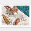 Two people with manicured hands and jewelry play dominoes on marble by a poolside, with a gold domino box and drink nearby in this sunlit scene—featured in the Poolside Dominoes Unframed Art Print by Shaynna Blaze x Urban Road.